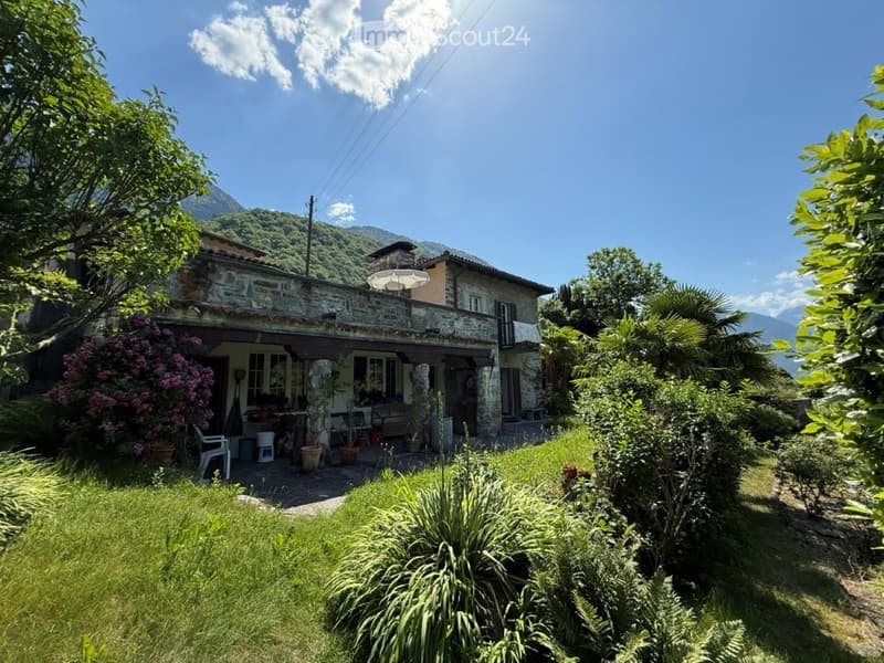 A rustic stone house with a tiled roof, surrounded by a well-maintained garden with various plants and trees. The house features a covered porch area with outdoor furniture and a scenic mountain view in the background.