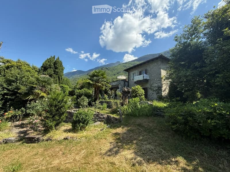 A rustic stone house surrounded by a lush garden with various plants, trees, and a scenic mountain view in the background.