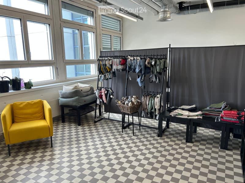 A modern, well-lit room with a checkered floor, large windows, a yellow sofa, a black table, a clothing rack, and a wicker basket with stuffed toys.