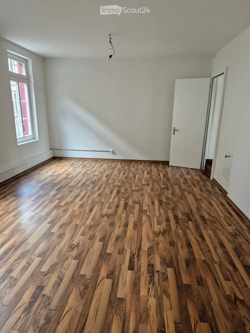 Empty room with wooden flooring, white walls, and a single window on the left side. A white door is visible on the right side.