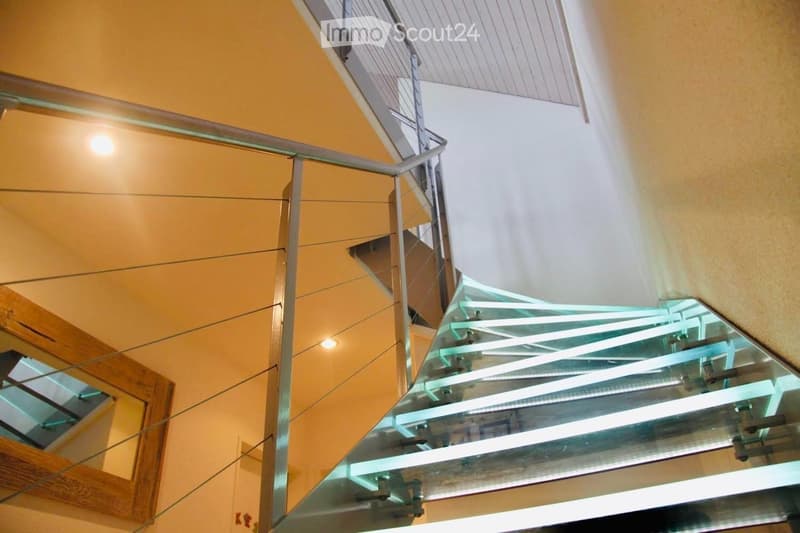 Modern glass staircase with metal railings, wooden handrails, and warm lighting, leading to an upper level.