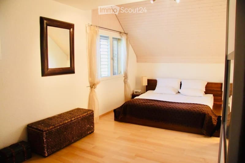 Cozy bedroom with wooden flooring, a large bed with white pillows and a dark brown duvet, a wicker storage box, a nightstand with a lamp, and a large mirror on the wall.