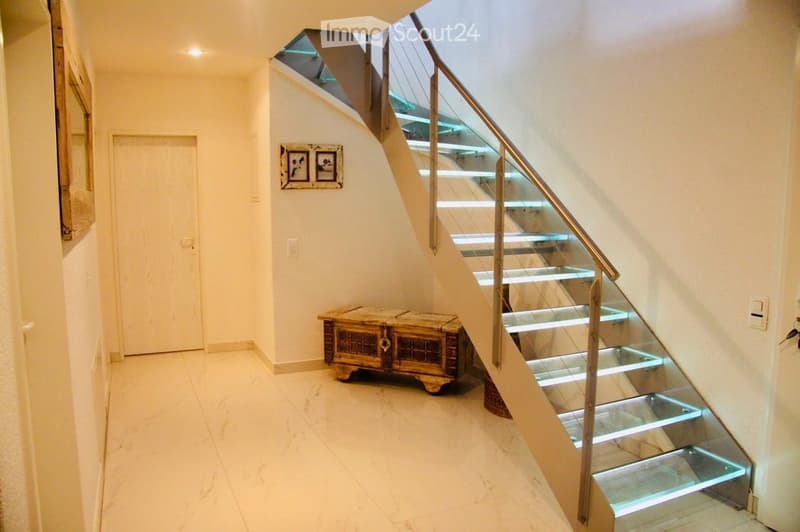 Modern hallway with a sleek staircase featuring glass steps and a metal railing, illuminated steps, a wooden chest, and a framed picture on the wall.