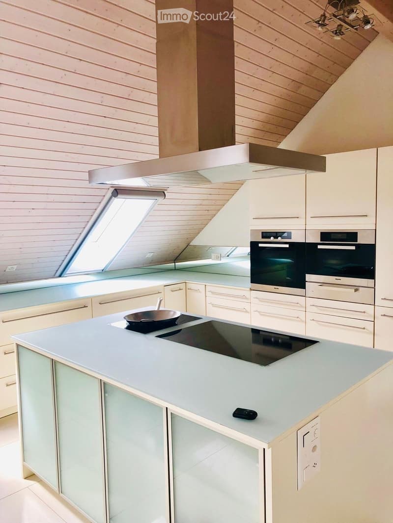 Modern kitchen with light-colored cabinets, an island with an induction cooktop, and built-in ovens. The island features a sleek, light blue countertop with a frypan and a remote control on it.
