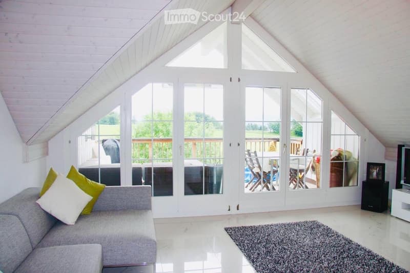 Bright, airy living space with a grey sofa, colorful pillows, a coffee table, and a TV. Large windows and sliding doors open to a patio with outdoor furniture.
