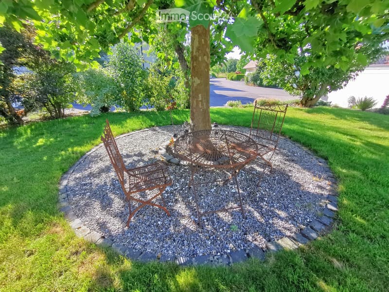 A small outdoor seating area with a round metal table and two matching chairs, surrounded by gravel and a tree, with a well-maintained lawn and garden in the background.