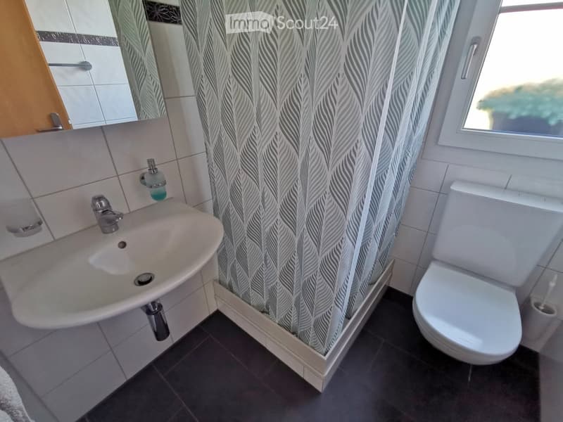 Modern bathroom with a white sink, chrome faucet, toilet, mirror, and a patterned shower curtain. The room features tiled walls and dark tiled flooring.