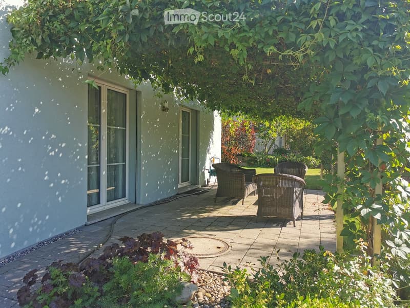 A sunny patio area with wicker chairs, surrounded by lush greenery and a well-maintained garden.