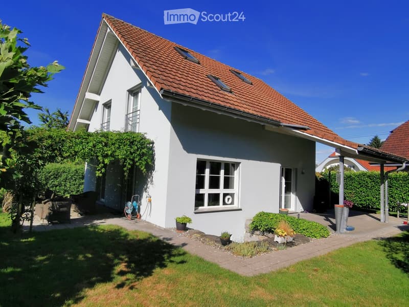 Single-story house with white walls, red-tiled roof, multiple windows, and a small balcony. The surrounding area includes a well-maintained lawn, garden beds, and a paved patio.