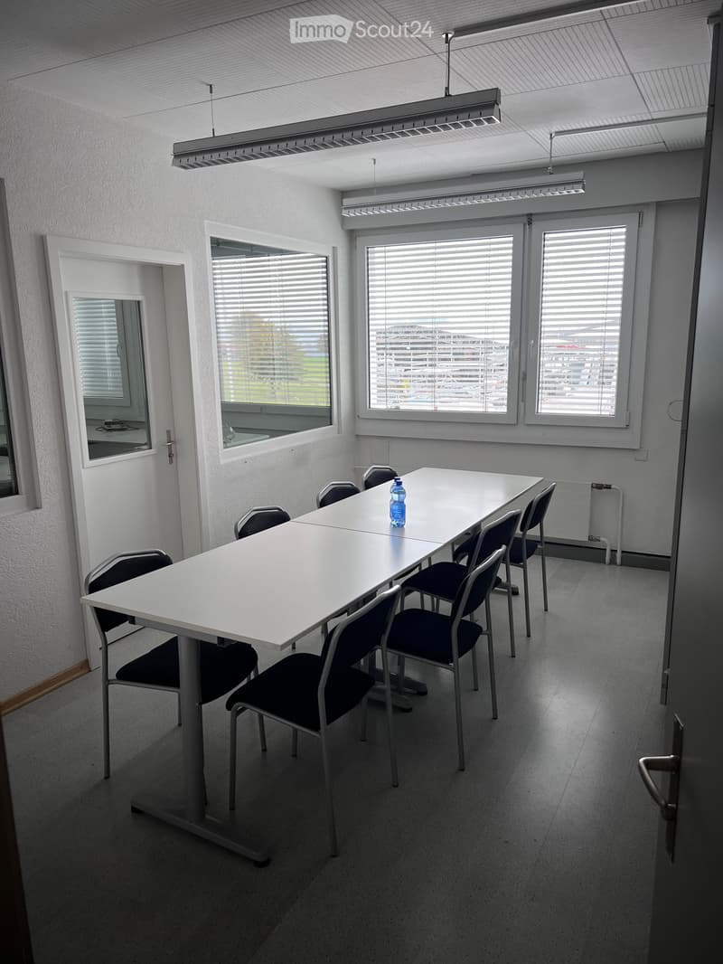 a large room with a long white table, and several chairs arranged around it, three windows with blinds, two doors, one of which leads to an outside area, a radiator, and a bottle on the table