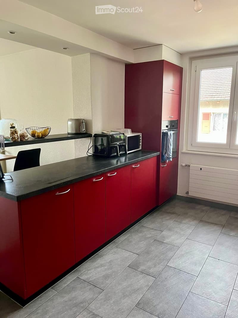 Modern kitchen with red cabinets, black countertop, and various appliances including an oven, microwave, toaster, and a chair.