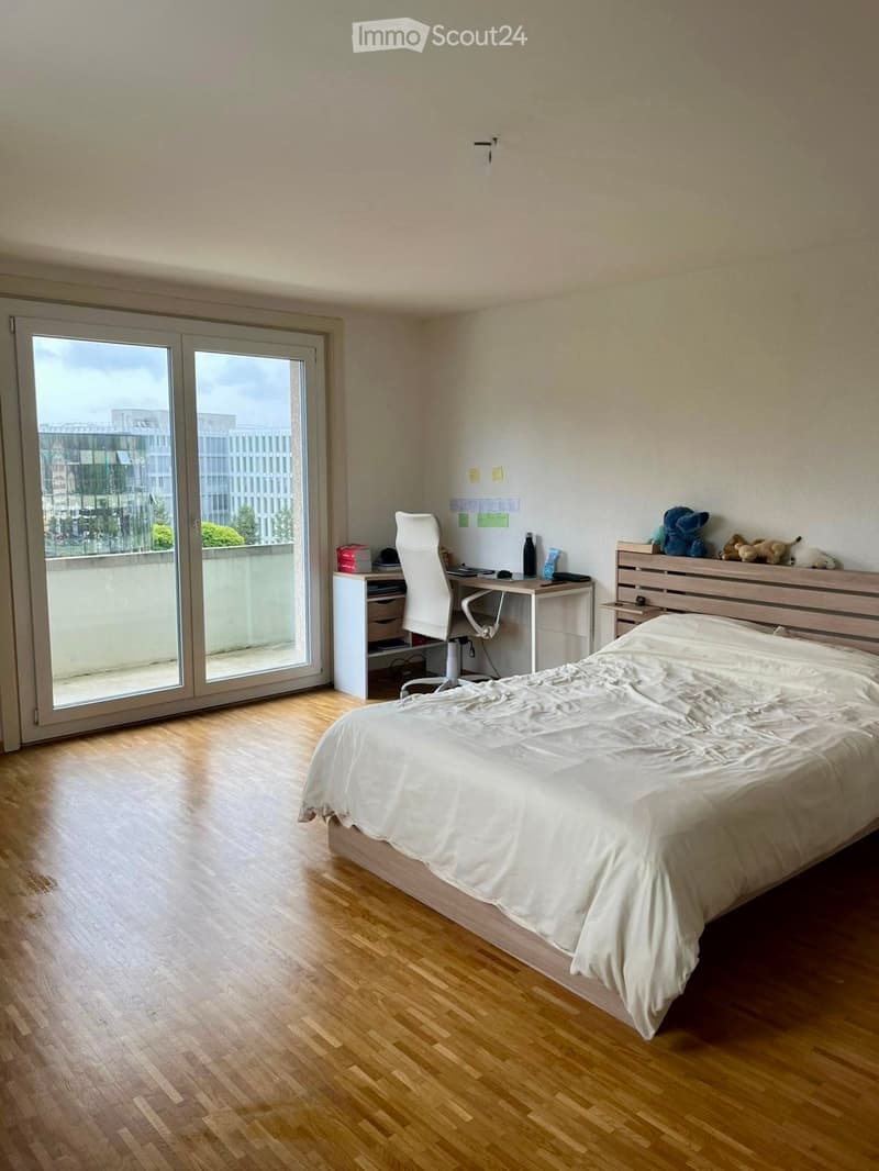 Bright bedroom with wooden flooring, large window, bed with white bedding, desk with chair, and storage unit.