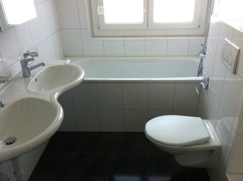 Modern bathroom with white tiles, a double sink, a bathtub, and a toilet. The room features a large window and sleek, contemporary fixtures.