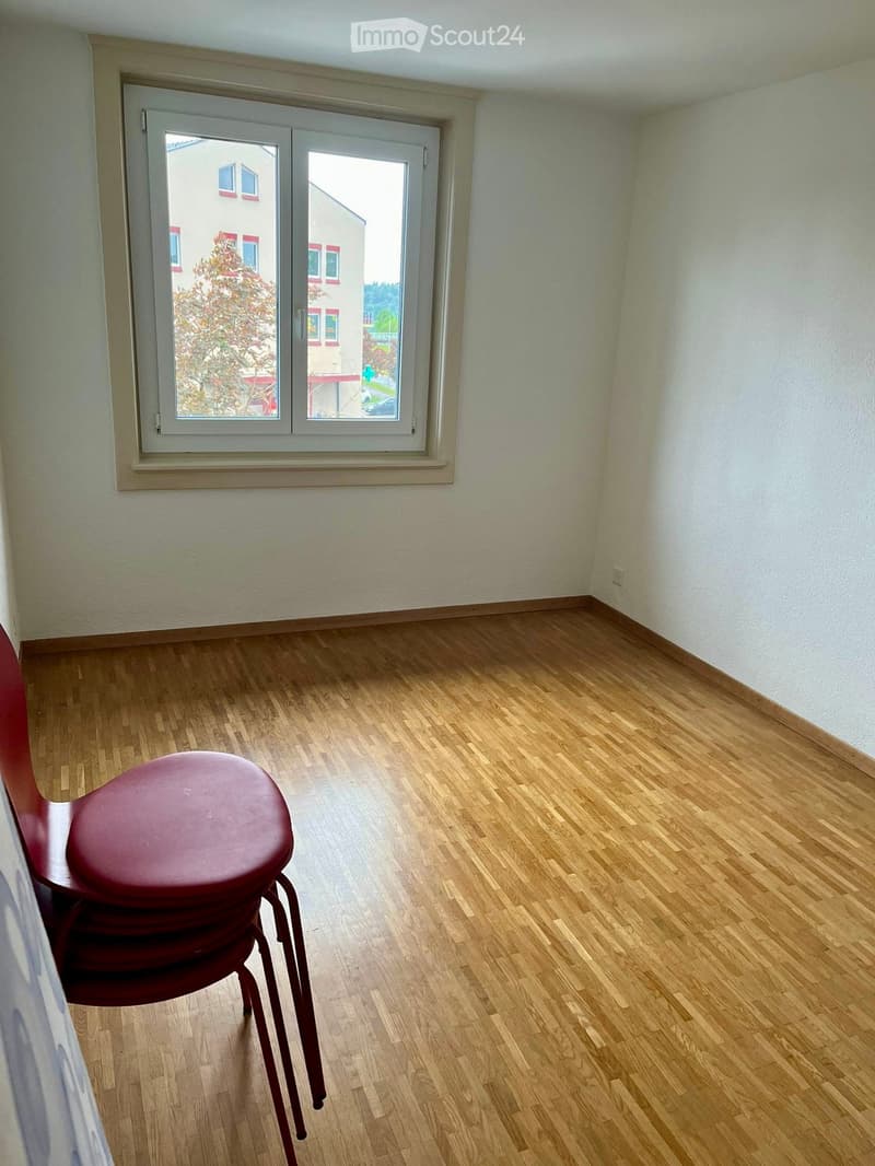 Empty room with wooden flooring, white walls, and a large window with a view of a neighboring building and tree. A red chair is placed near the window.