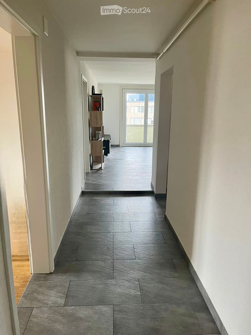 Clean, modern hallway with grey tiled flooring and white walls, leading to a glass door at the end.