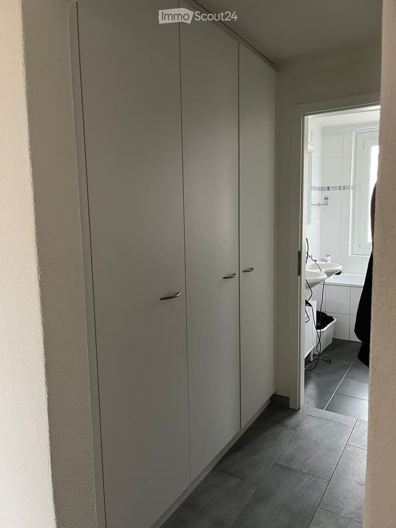 A narrow hallway with grey tiled flooring and white walls, leading to a small bathroom with a sink, bathtub, and window.