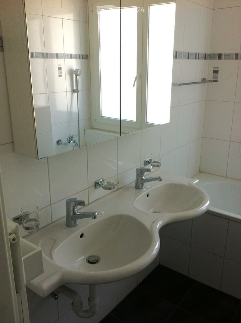 Modern bathroom with double sink, chrome faucets, large mirror, bathtub, and glass on counter.