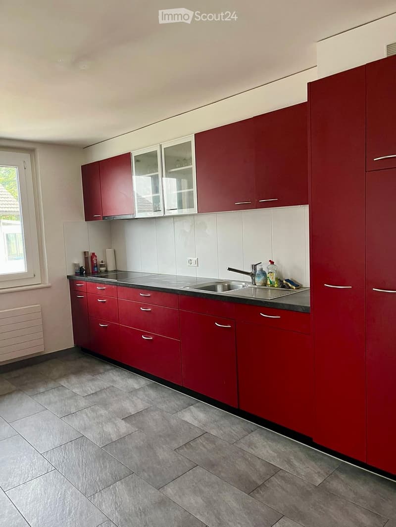Modern kitchen with red cabinets, white tiles, and a stainless steel sink. Includes a window, radiator, and various small items on the counter.