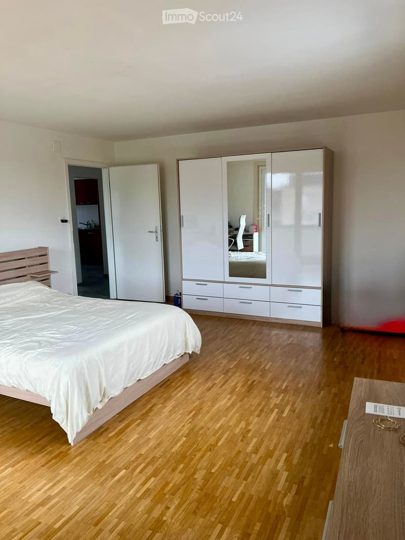 Modern bedroom with wooden flooring, a large bed with white bedding, a white wardrobe with mirrored doors, and a small table with decorative items.