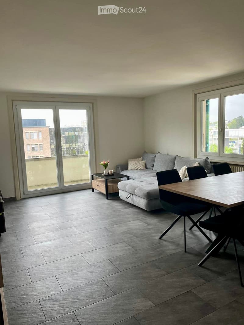 Modern living and dining area with grey sectional sofa, coffee table, dining table, and chairs. Large windows and sliding glass door provide natural light.