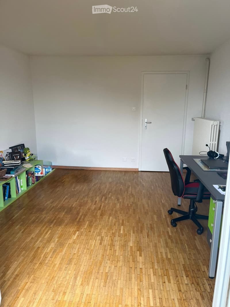 A furnished room with wooden flooring, a desk with a chair, a bookshelf, and a radiator on the wall.