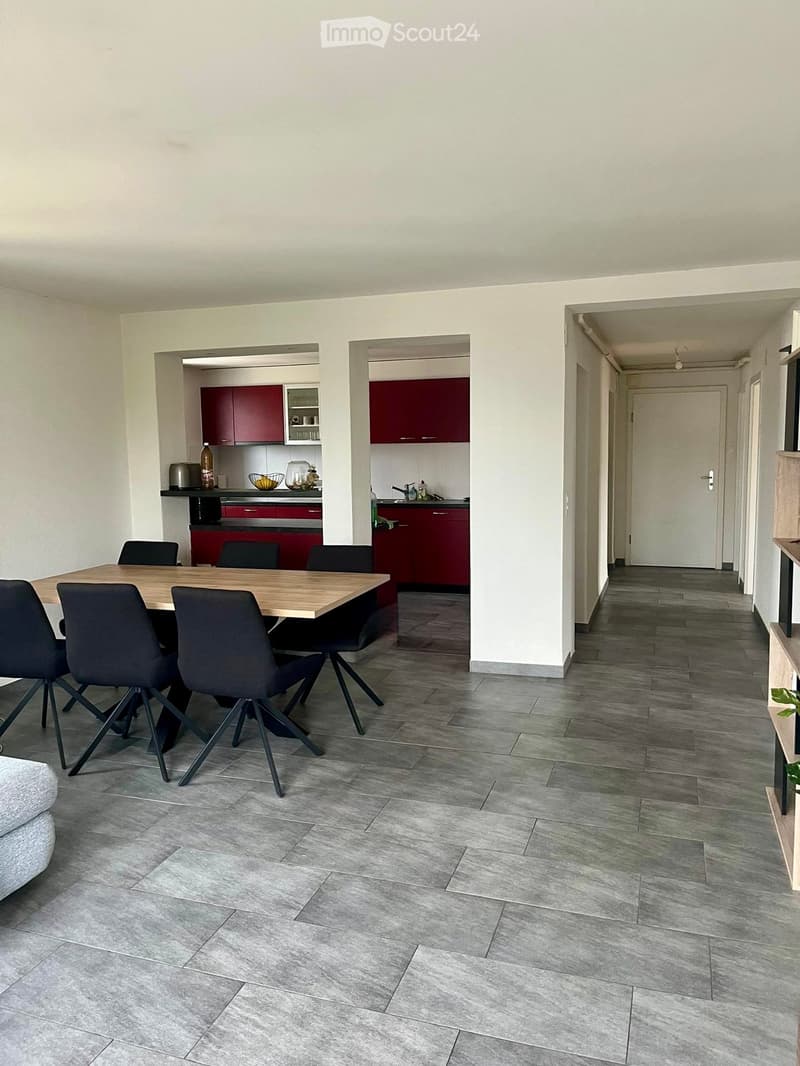 Open-plan living and dining area with grey tiled flooring, a wooden dining table surrounded by black chairs, and a modern kitchen with red cabinets and stainless steel appliances.