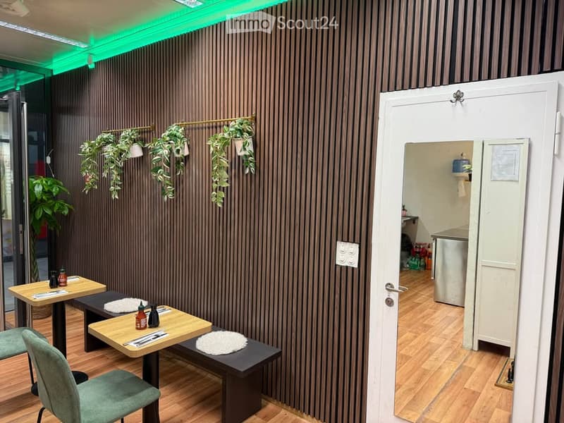 A modern, stylish room with wooden flooring, dark vertical wall panels, and a mix of seating and tables. Potted plants are displayed on the wall and in corners, and a small kitchen area is visible through an open doorway.