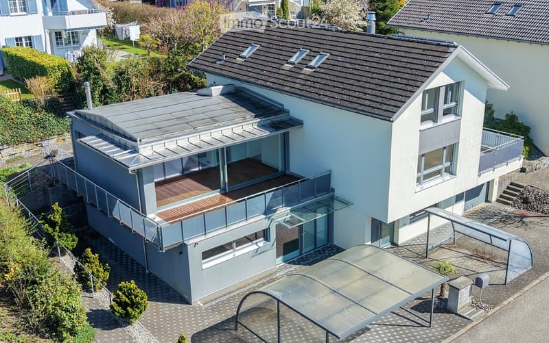 Modern two-story house with a flat roof, large windows, and a spacious balcony area. The house features a sleek design with a combination of light-colored walls and dark roofing. The surrounding area includes a paved driveway, small garden, and neighborin