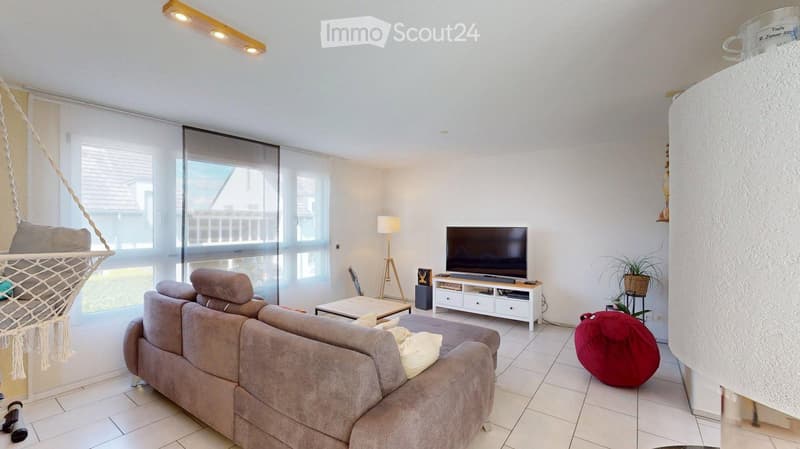 Modern living room with large windows, grey sectional sofa, coffee table, TV, floor lamp, and a hanging hammock.