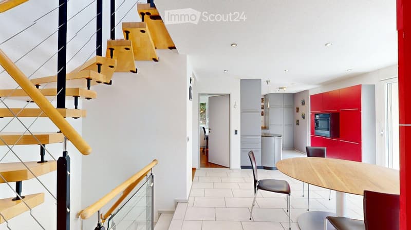 Modern interior with a wooden staircase, a round wooden dining table with chairs, red kitchen cabinets, and a built-in oven.
