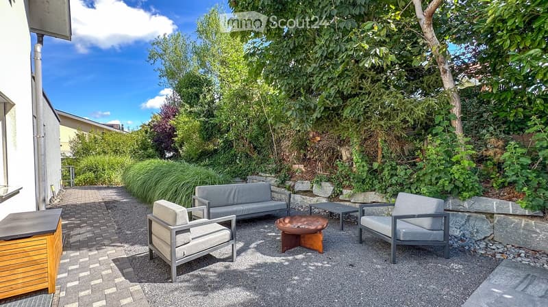 A well-maintained garden patio with a grey sofa, two grey chairs, a small table, and a decorative bowl in the center. The area is surrounded by lush greenery and has a paved pathway leading to the house.