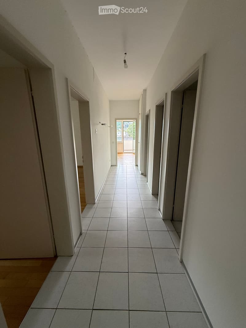 Empty hallway with tiled floor, white walls, and several doors leading to other rooms. Natural light comes from an open doorway at the end.