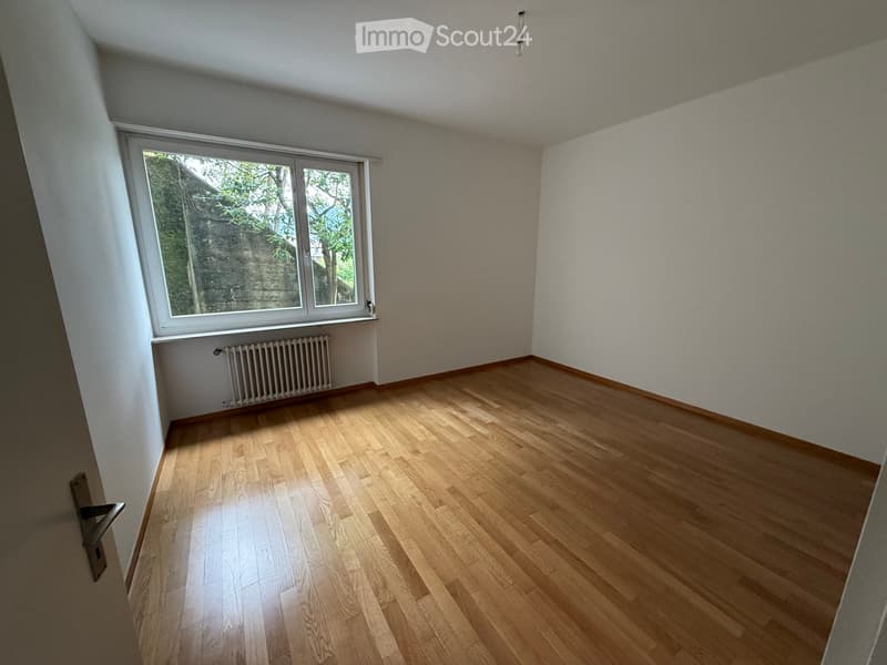 Empty room with wooden flooring, white walls, and a large window with a view of greenery. A radiator is visible below the window.