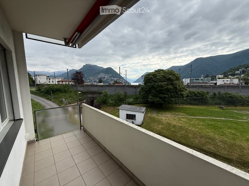 Balcony with tiled floor, glass railing, overlooking a grassy area with a small shed and distant mountains.