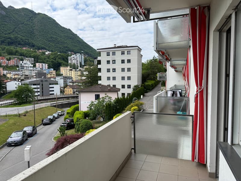 Balcony with tiled floor, glass railing, and red striped curtains, overlooking a street with parked cars and a mountainous backdrop.