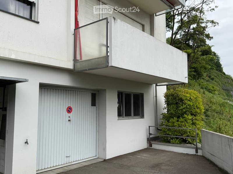 Modern two-story building with white exterior, garage, and balcony overlooking a green hillside.