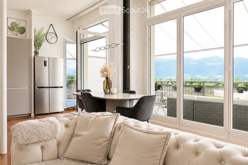 Bright, modern living and dining area with large windows offering a scenic mountain view. Features a white tufted sofa, coffee table, dining table with chairs, stainless steel refrigerator, and decorative elements like a clock and vase with dried flowers.