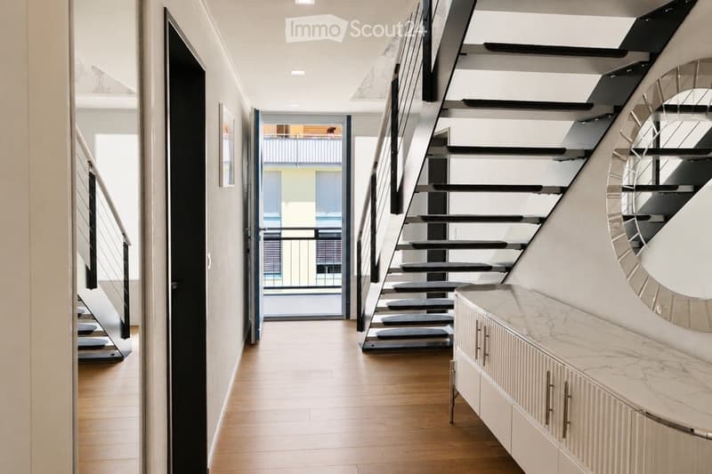 Modern hallway with wooden flooring, sleek staircase, and a large mirror on the wall. Features a door leading to a balcony area.