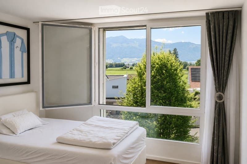 Bright bedroom with large windows offering a scenic mountain view, featuring a neatly made bed with white linens and pillows, a framed picture on the wall, and grey curtains.