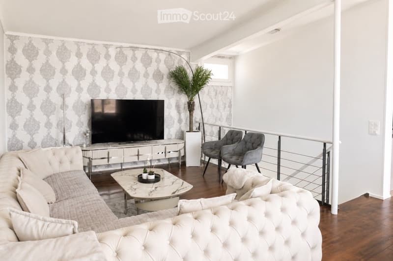 Modern living room with a grey sofa, coffee table, armchairs, ottoman, floor lamp, television, and a decorative wall. The room features a balcony with a railing and a potted plant.