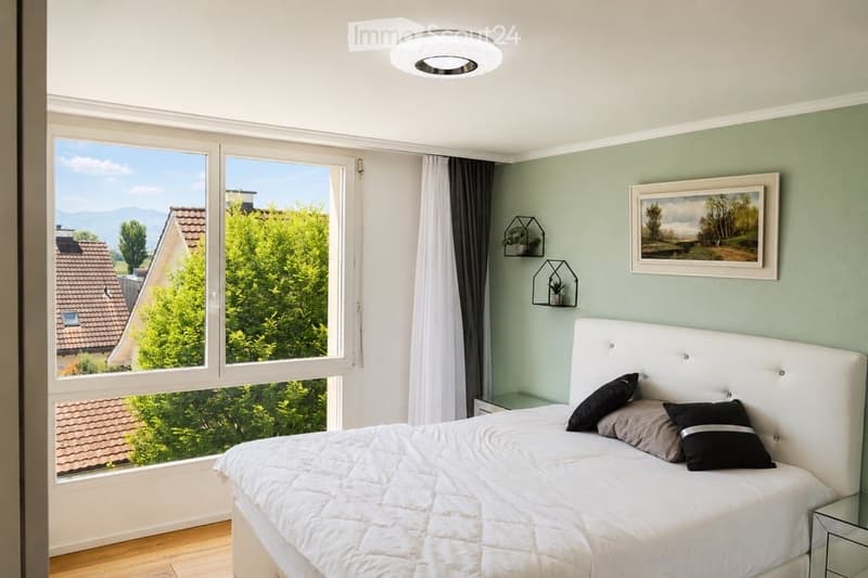Modern bedroom with a large bed, white bedding, multiple pillows, a nightstand, and a view of greenery outside. The room features light green walls, a ceiling light, and decorative wall-mounted shelves with small plants.
