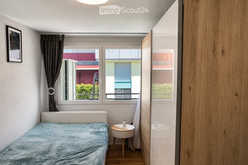 Cozy bedroom with a single bed, nightstand, small stool, and a picture frame on the wall. The room features large windows with curtains and a view of the outdoor area.