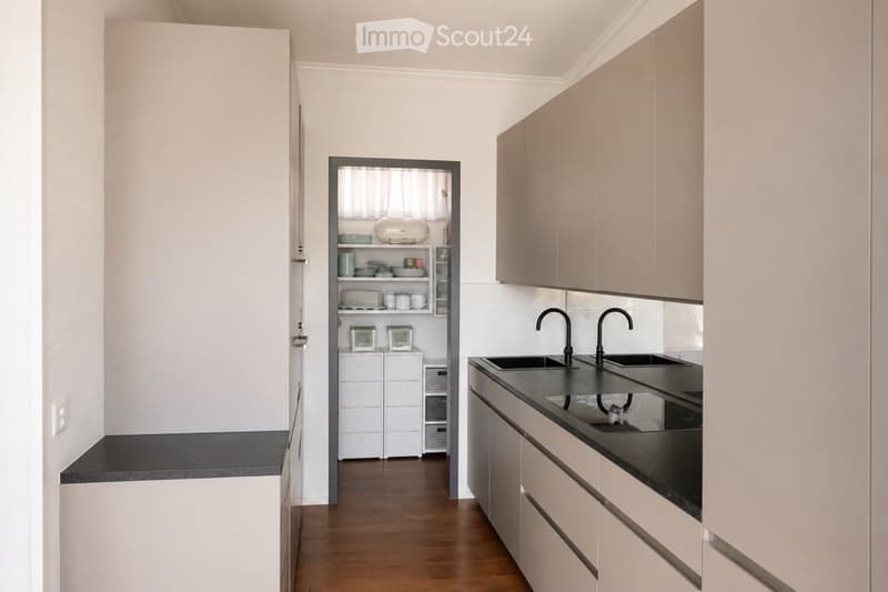 A modern hallway leading to a kitchen area, featuring light-colored cabinets, a black countertop, and a double sink with a modern faucet.