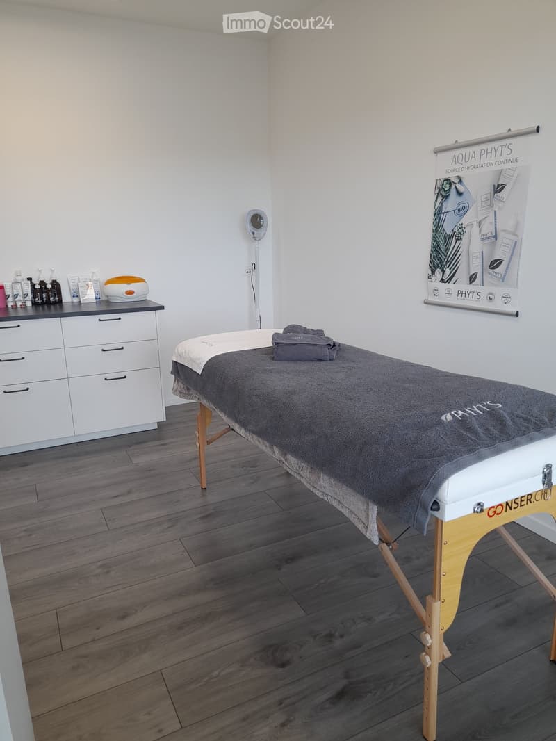 A clean, well-lit massage room with a wooden massage table, a white cabinet with various cosmetic products, and a mirror on the wall.