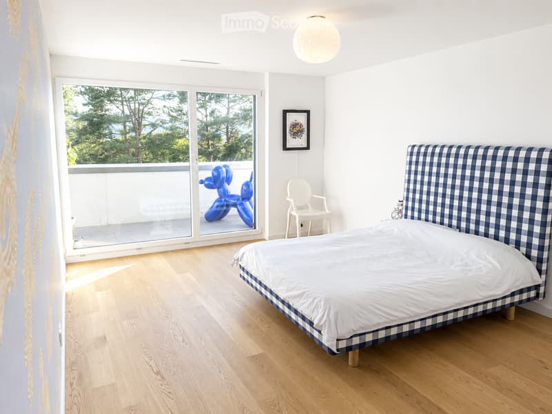 Modern bedroom with a large bed, white bedding, blue and white checkered headboard, a white chair, and a view of greenery through large windows.
