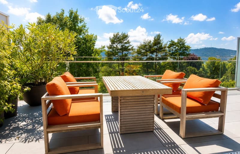 A modern outdoor terrace with wooden chairs, a table, and orange cushions, surrounded by greenery and a scenic mountain view.