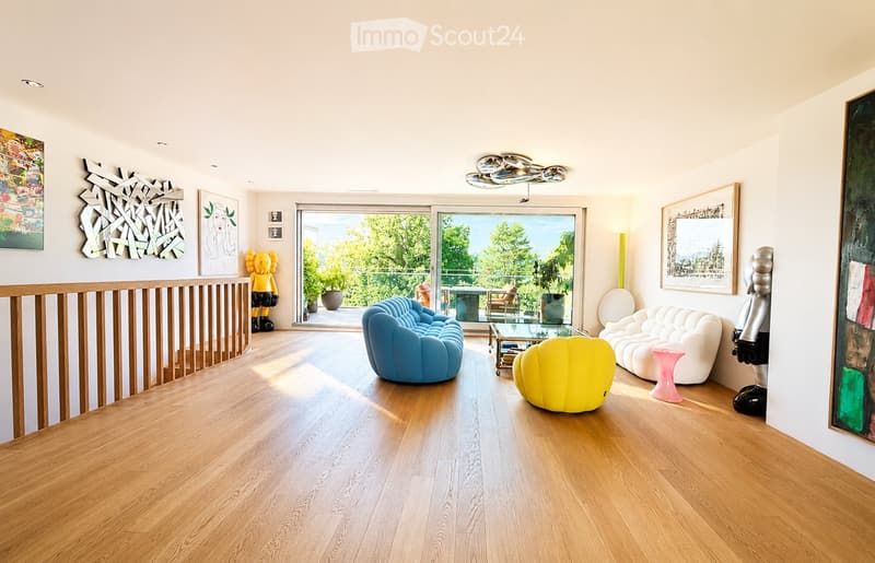 Modern living room with light wooden flooring, large windows offering a view of greenery, and contemporary furniture including a blue sofa, white sofa, yellow chair, glass coffee table, and decorative art pieces.