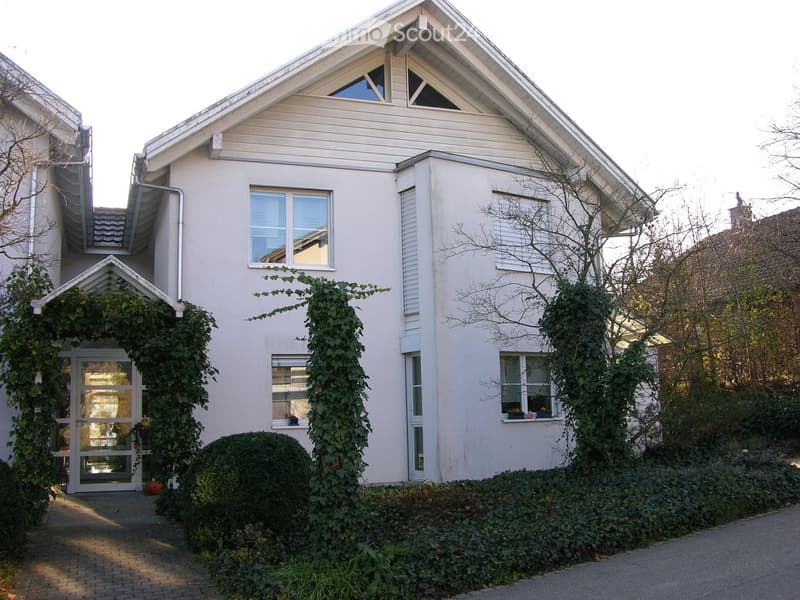 Single-story house with light-colored exterior, multiple windows, and a covered entrance with climbing plants.