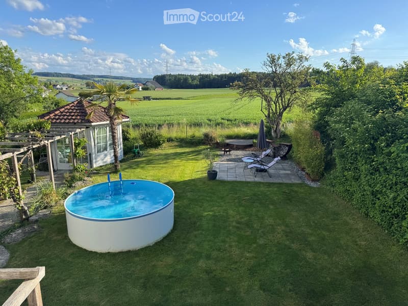 Garden with a small round swimming pool, lounge chairs, a pergola, and a view of green fields and trees.