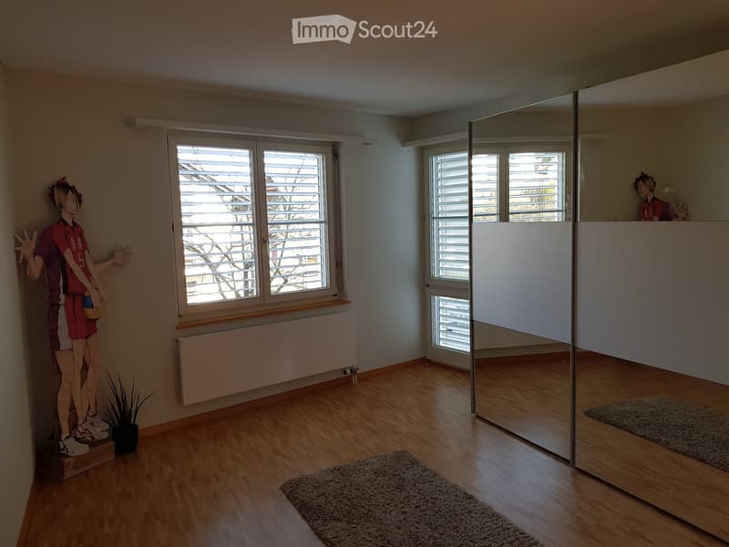 A modern, well-lit room with wooden flooring, large windows with blinds, a radiator, a rug, a small plant, and a large mirror. The room features a large anime-style cardboard cutout of a character near the window.